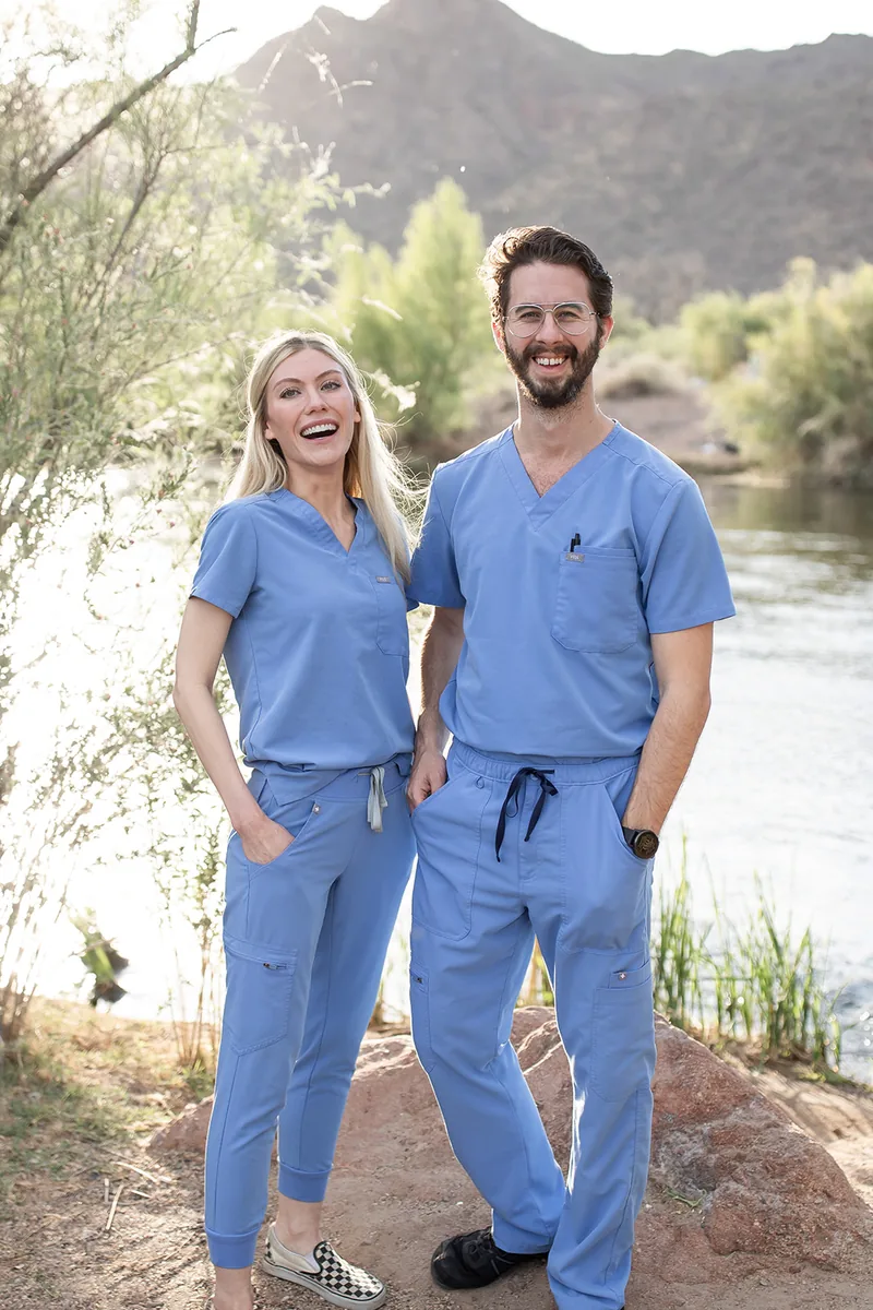 Dr. Kaitlyn Myers and Dr. Drew Timmermans, co-founders of Regenerative Performance, standing side by side in light blue scrubs beside the water with a desert mountain behind them
